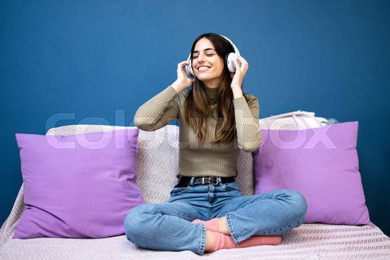 Cheerful young woman listening to music and smiling sitting on couch in livinig room. Happy young girl having fun at home.