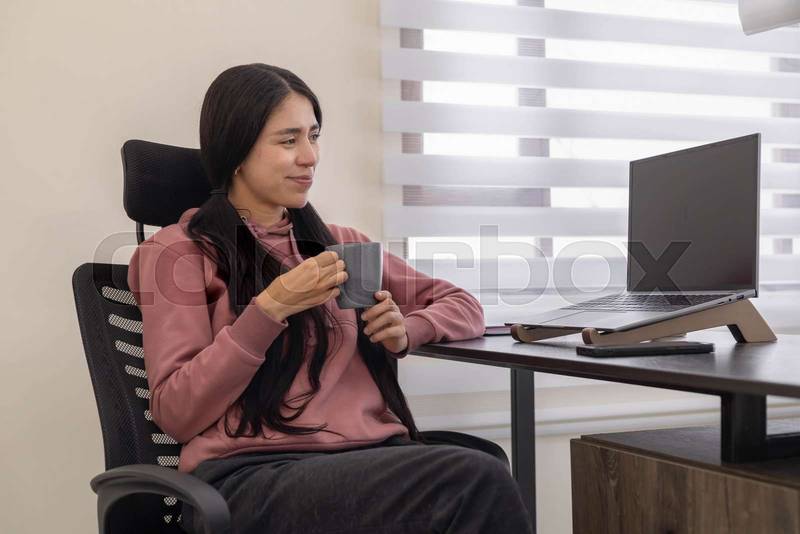 sits at her office desk with a laptop, holding a cup of coffee, taking a short pause