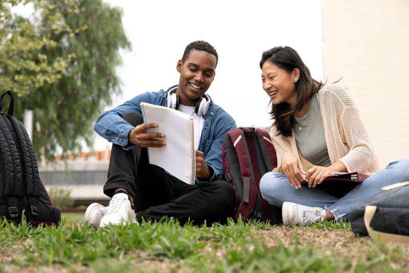 Diverse couple of students sitting outside reading documents.Two young multiracial students looking at notes sitting on the floor campus.