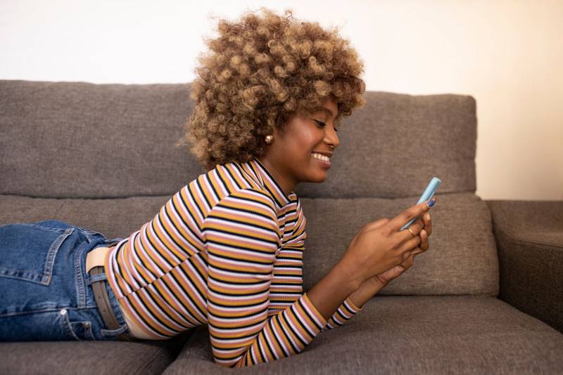 Smiling beautiful girl lying on a couch at home texting a with her smartphone. Happy young female using a mobile phone while relaxing at sofa.