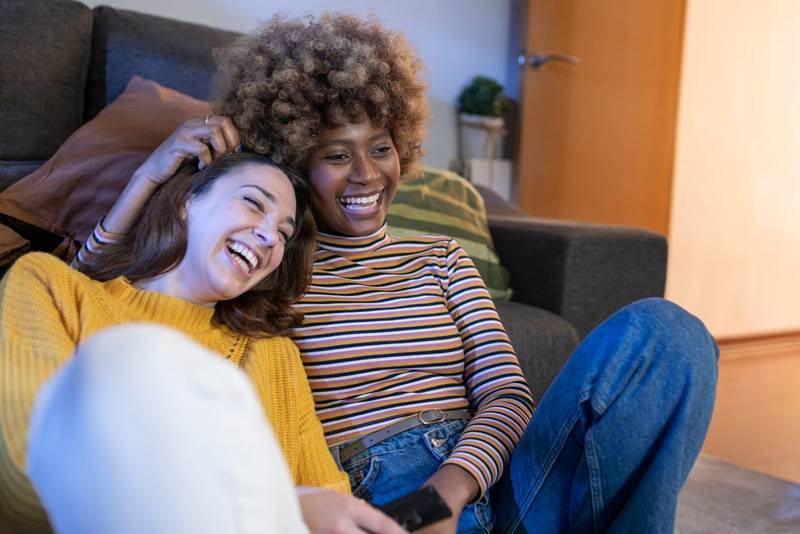 Happy lesbian couple watching television relaxed in the floor at home. Two women laughing while watching comedy at home on tv.