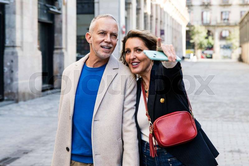 Carefree middle aged couple looking for touristic location pointing and holding smartphone. Happy senior man and woman sightseeing and exploring a city in a trip.