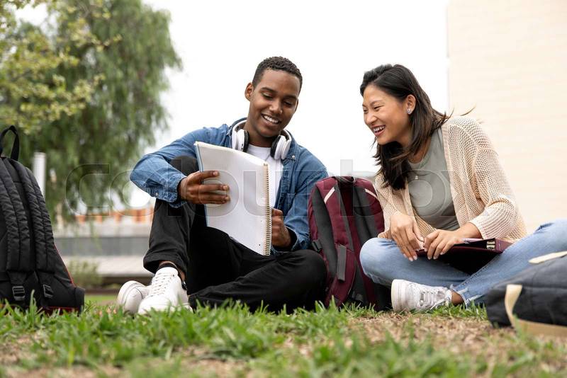 Diverse couple of students sitting outside reading documents.Two young multiracial students looking at notes sitting on the floor campus.