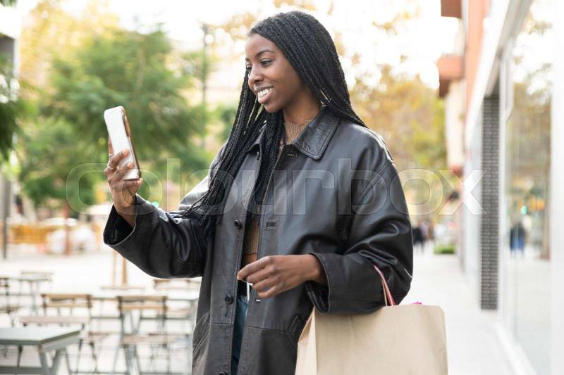 Happy young woman taking a videocall on phone in the street holding a shopping bag. Happy female using a smartphone in the street