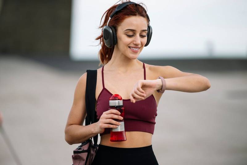 Young fitness female drinking water outside wearing headphones. Athletic woman using a bottle and a gym bag ready for training and workout.