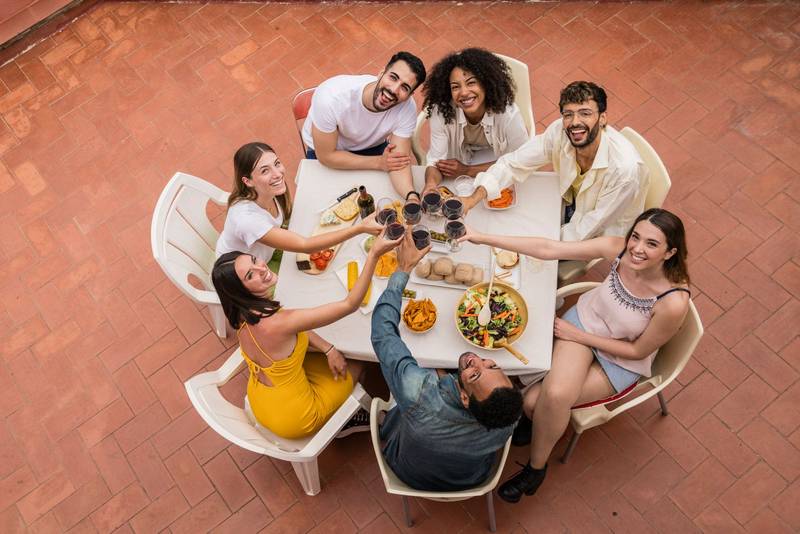 Top view of a group of happy young friends toasting with glasses in a terrace. Carefree and cheerful people sitting in a patio having dinner and drinking wine.