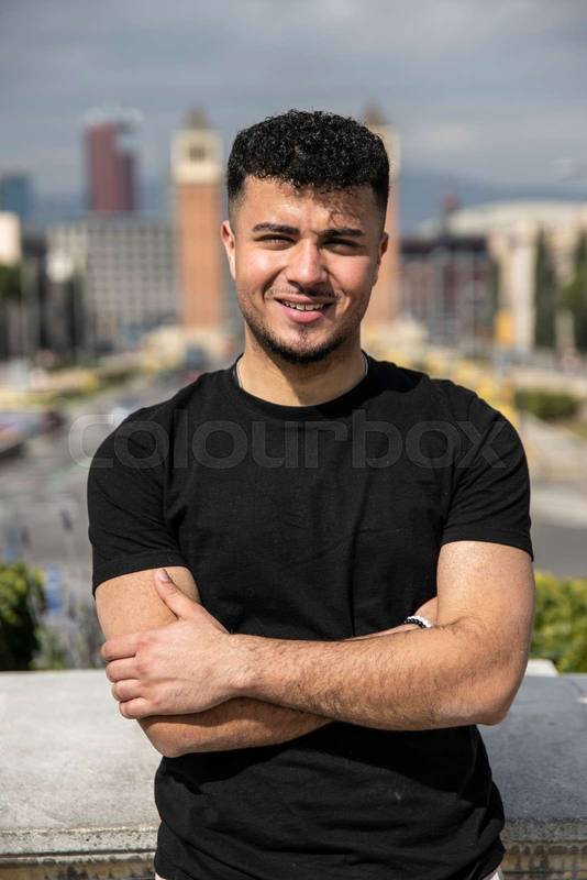 Close up of a young handsome guy smiling relaxed and looking at camera in the street with arms crossed. Confident happy man relaxed and staring at camera standing with arms folded outdoors.