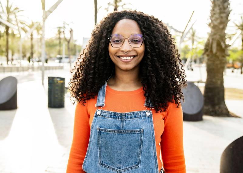 Portrait of beautiful happy curly hair young woman smiling and looking at camera in the street. Gorgeous cheerful female confident and satisfied standing outside 