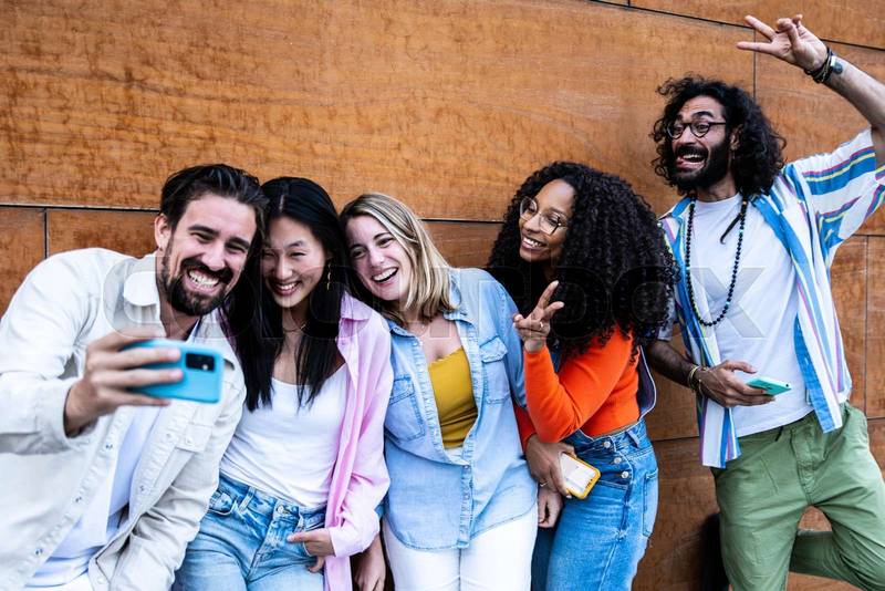 Multiethnic group of friends taking a selfie while standing on a wooden wall in the street. Diverse young people smiling and taking a picture outside in a city with a brown background.