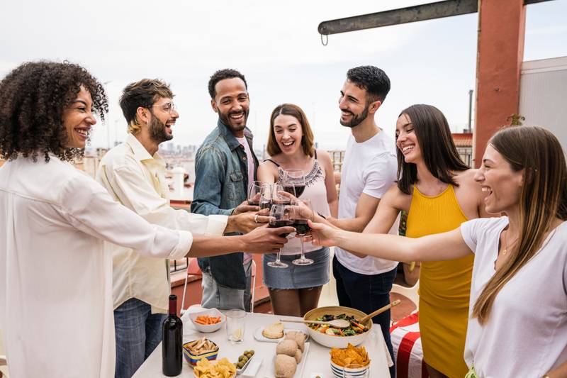 Happy group of friends toasting with wine glasses standing around a table in a terrace. Diverse group of cheerful young people drinking and celebrating in a rooftop dinner.