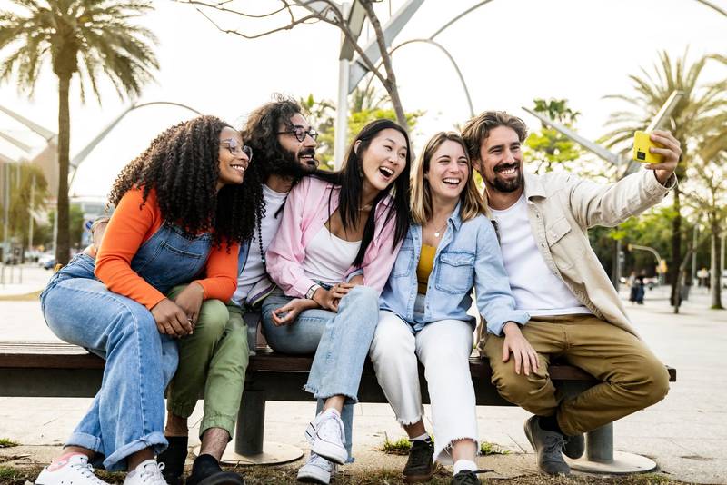 Diverse group of friends taking a selfie in the street sitting on a bench. Cheerful multiracial group of young hipsters taking a picture outside in a park bench.