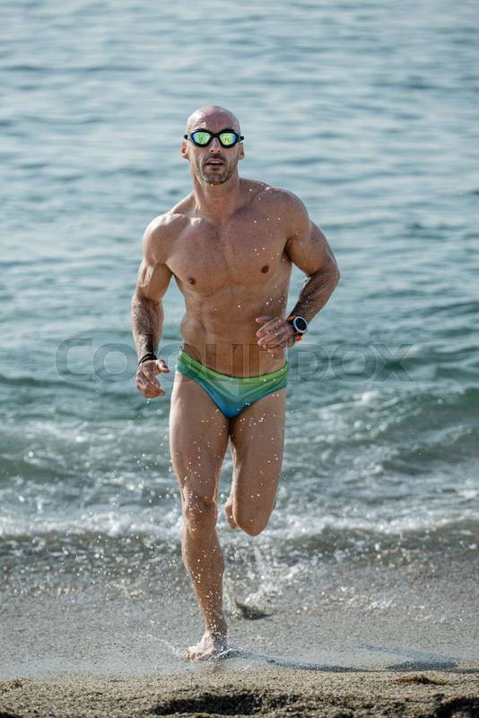 Sexy muscular man in colorful speedo running out of the sea looking at camera. Professional male athlete getting out of the water wearing swim glasses at the beach.
