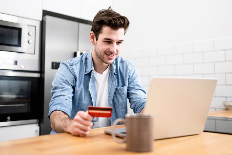 Man sitting in front of the computer holding a credit card at home - Young adult doing a online payment with his laptop at kitchen - business, technology concept