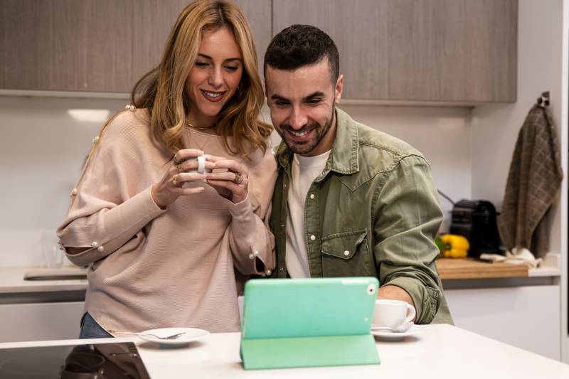 Young adult couple using together a tablet from home in a modern kitchen. Happy joyful man and woman on a video call having a coffee cup.