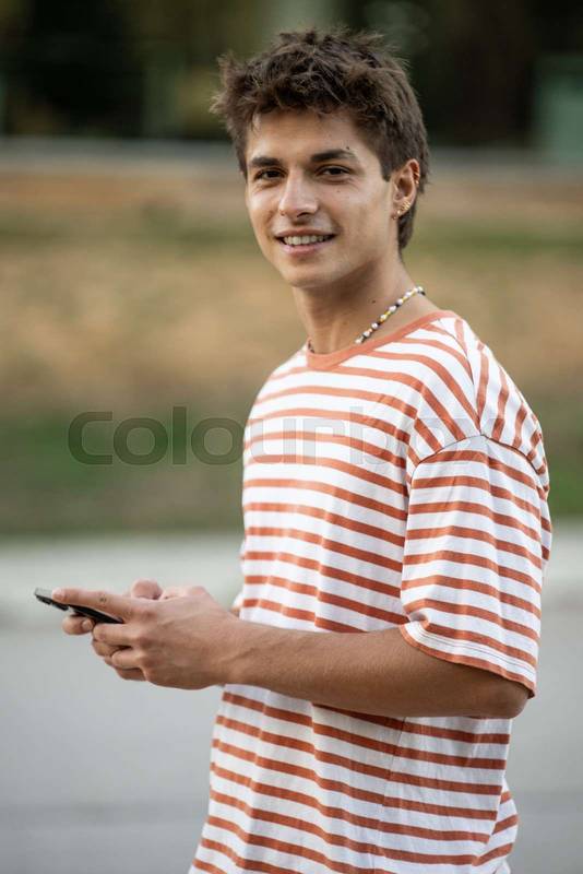 Confident young guy looking at camera and texting with smartphone.Satisfied teenager standing relaxed outside and smiling using his phone.