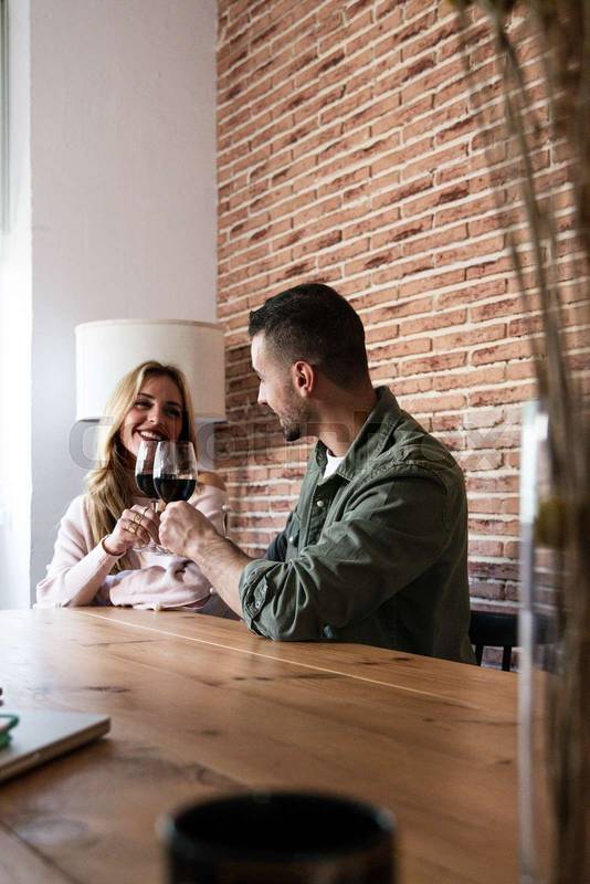 Affectionate couple smiling in a date drinking alcohol sitting at table in a house. Joyful woman and man drinking wine together in a living room with glasses.