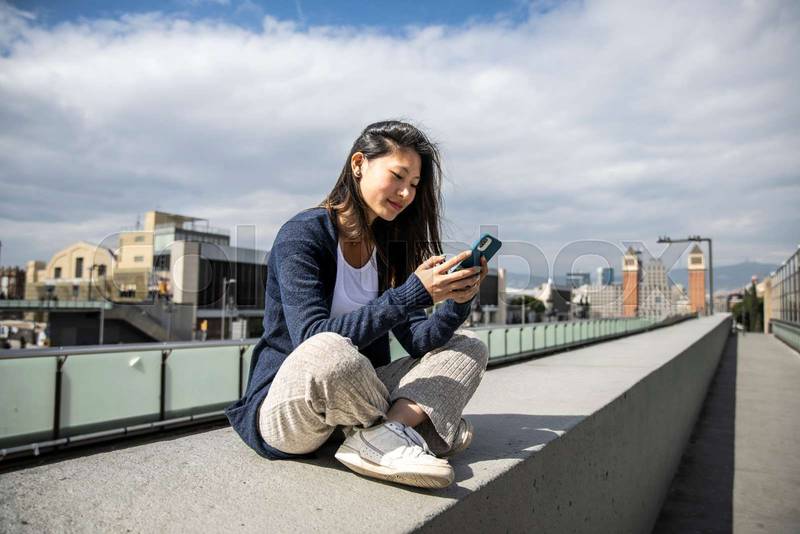 Young beautiful girl sitting on bank relaxed and texting with her phone. Satisfied woman using an smartphone sitting in the street.