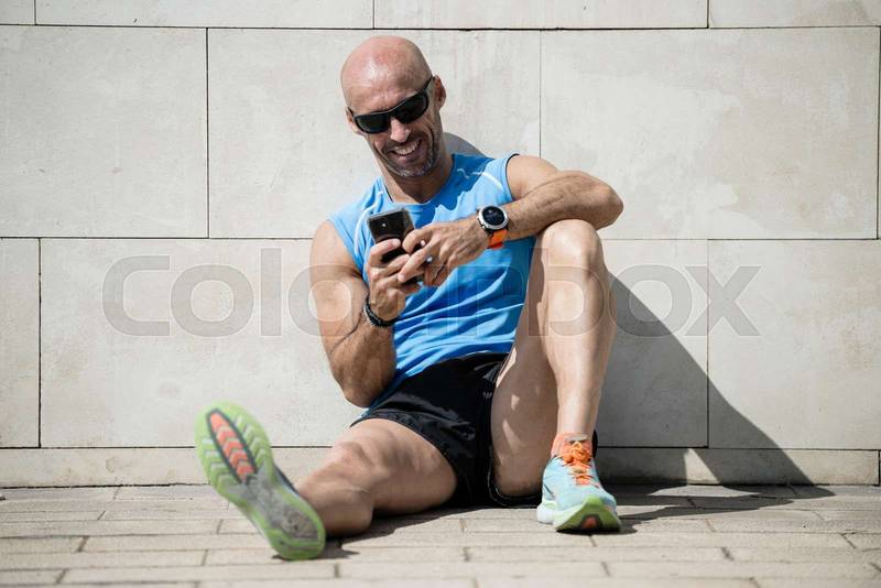 Fit happy man checking his phone sitting on the street floor. Relaxed smiling athletic runner male watching his smartphone resting against a wall in the city.