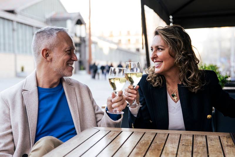 Lovely mid adult couple toasting with wine while sitting in a terrace restaurant. Romantic date of a middle age woman and man drinking with glasses at a bar terrace looking each other.