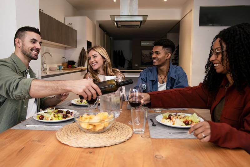 Diverse group of happy friends having party diner at home. Smiling people eating and drinking on modern apartment.