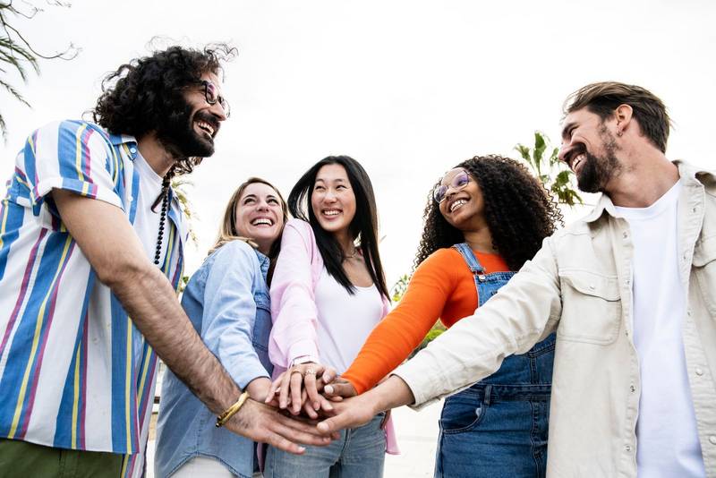 Diverse group of cheerful millennial friends standing together stacking hands in the street. Cheerful multiethnic young carefree friends laughing and having fun outside joining hands in the center.