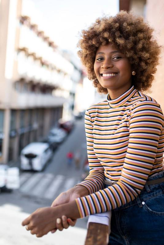 Portrait of smiling woman standing in a balcony with city background. Girl staring at camera with a smile in a high window.
