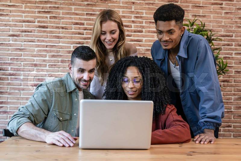 Diverse group of young adult friends working sharing a computer in a modern apartment. University students using laptop together.