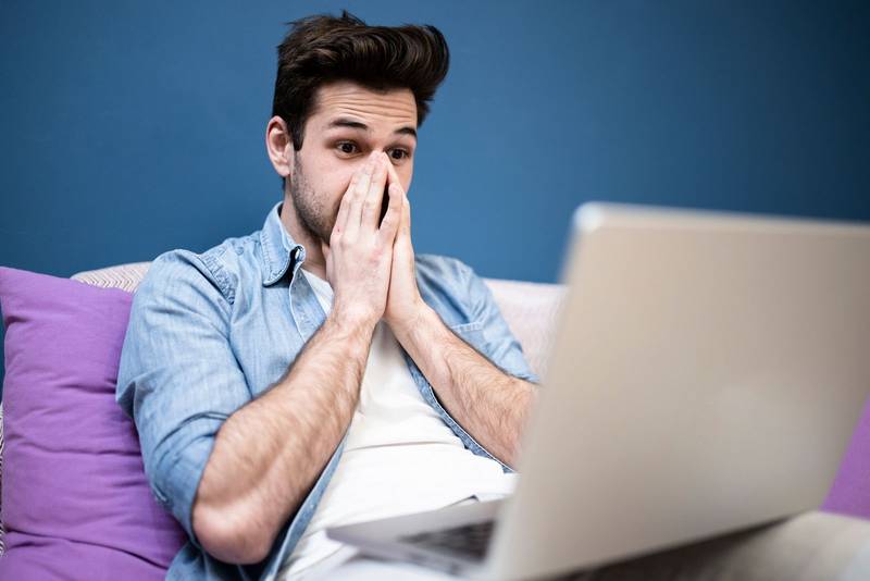 Excited handsome young man looking at laptop screen sitting on cozy sofa at home. Student reading amazing news on computer. Technology concept