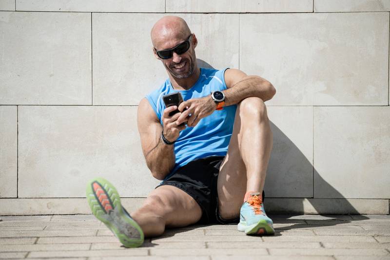 Fit happy man checking his phone sitting on the street floor. Relaxed smiling athletic runner male watching his smartphone resting against a wall in the city.