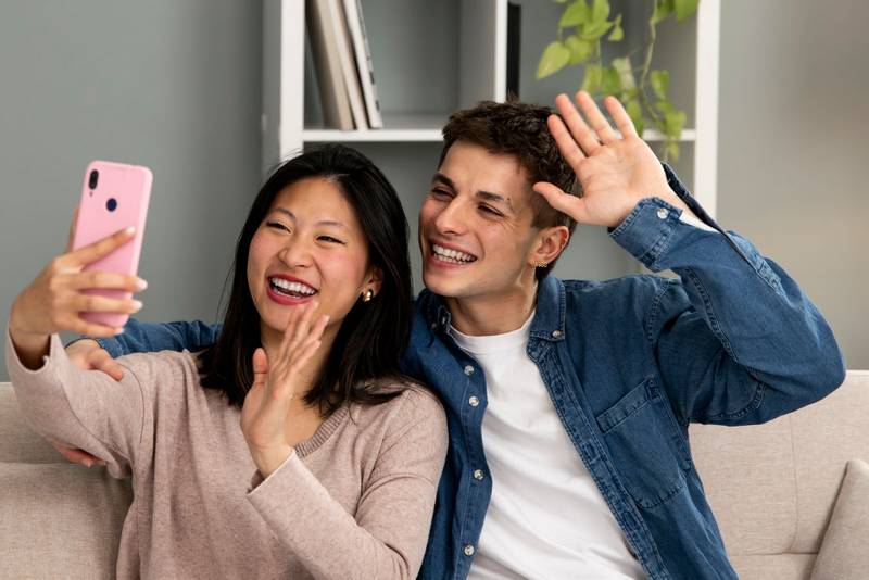 Diverse young couple on the sofa in their living room taking a selfie waving. Cheerful man and woman laughing in a video call on smart phone.