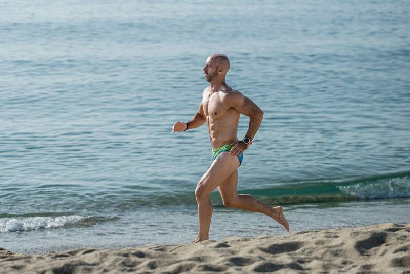 Athletic muscular man in swimwear running next to the sea. Professional male athlete training in speedo at the beach.