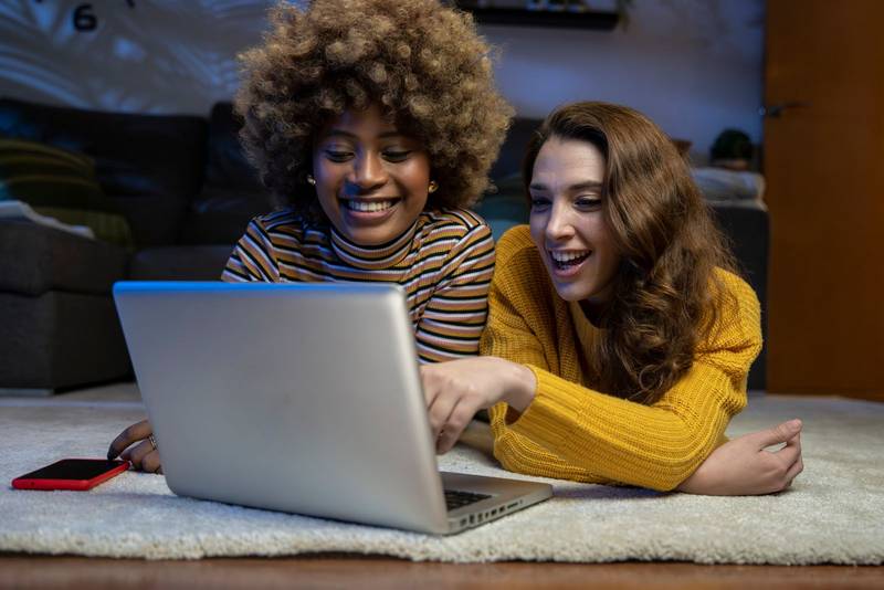 Two happy woman laughing at computer screen lying on the floor. Smiling lesbian couple lying on a carpet and using a laptop in a living room.