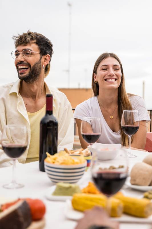 Happy young couple laughing sitting on a terrace with drinks and food. Cheerful man having fun with beautiful and relaxed lady in a rooftop dinner party.