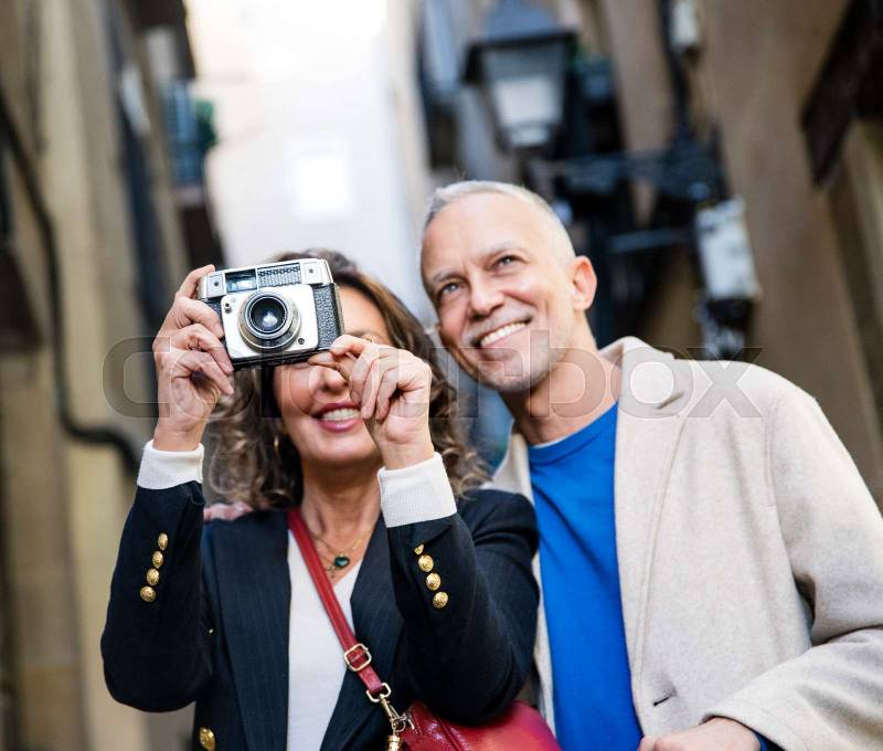 Senior tourist couple taking picture on vintage camera in old town. Mid adult woman taking photography in city street with her husband during a romantic trip.