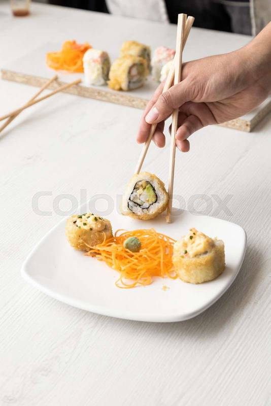 person's hand holding with chopsticks a sushi roll from a cardboard box, behind a plate with other ordered rolls