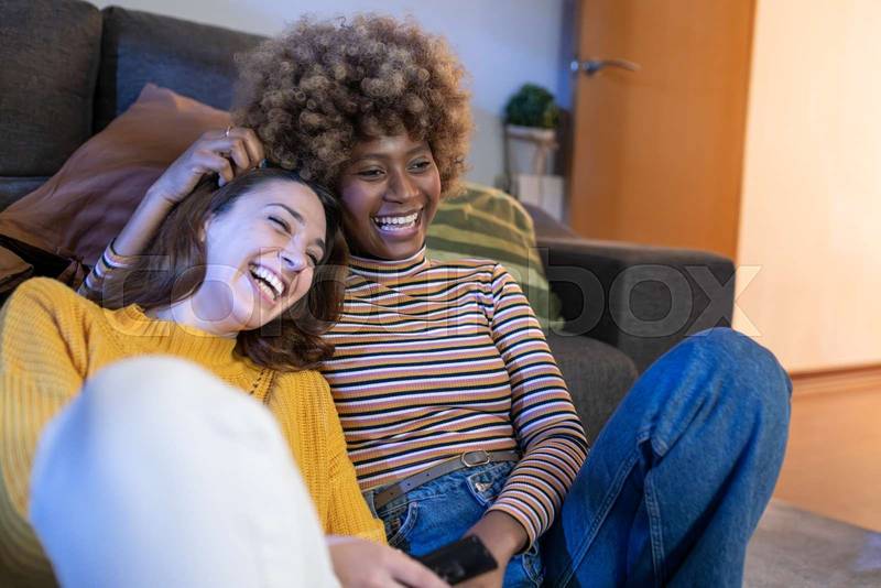 Happy lesbian couple watching television relaxed in the floor at home. Two women laughing while watching comedy at home on tv.