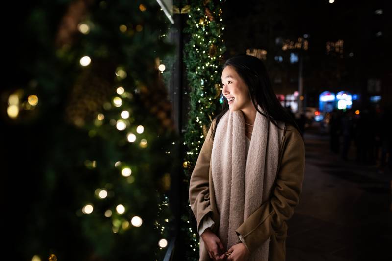 Hopeful young girl looking at shop window in a winter night. Smiling female holding bags with Christmas decoration shop.