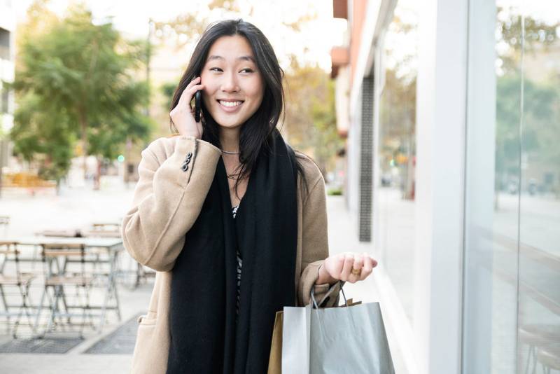 smiling young asian woman talking on a phone and holding shopping bags. happy female with a cellular in the city. Beauty, fashion, concept