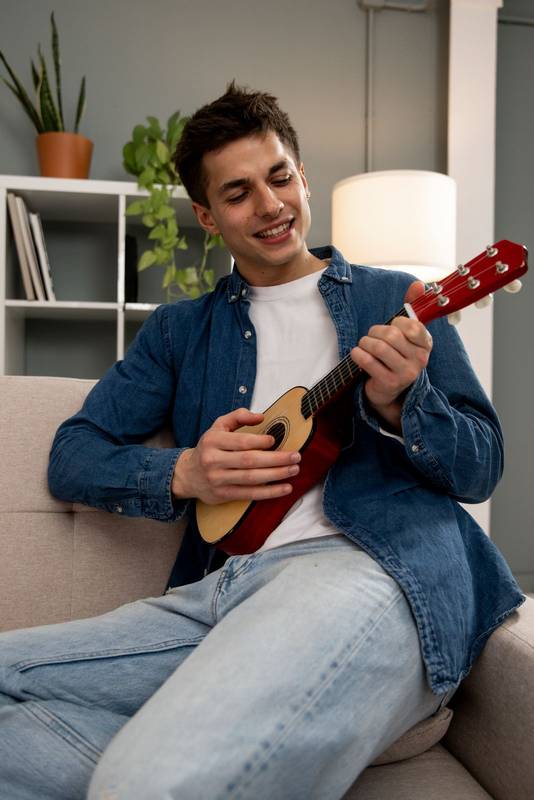 Young adult carefree male learning to play guitar on couch at living room. Young relaxed man playing music with ukelele sitting on sofa at home. 