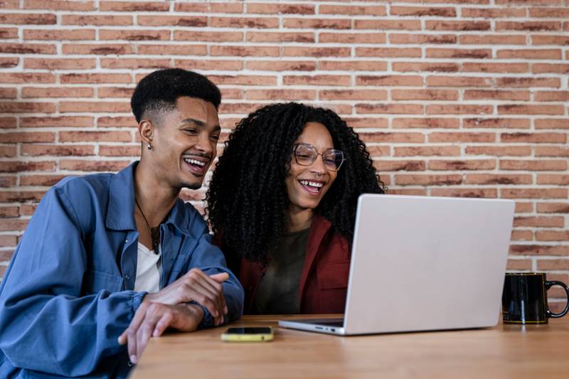 Diverse couple at home working online using laptop. Smiling man and woman young adult using laptop together while sitting with a mug. 
