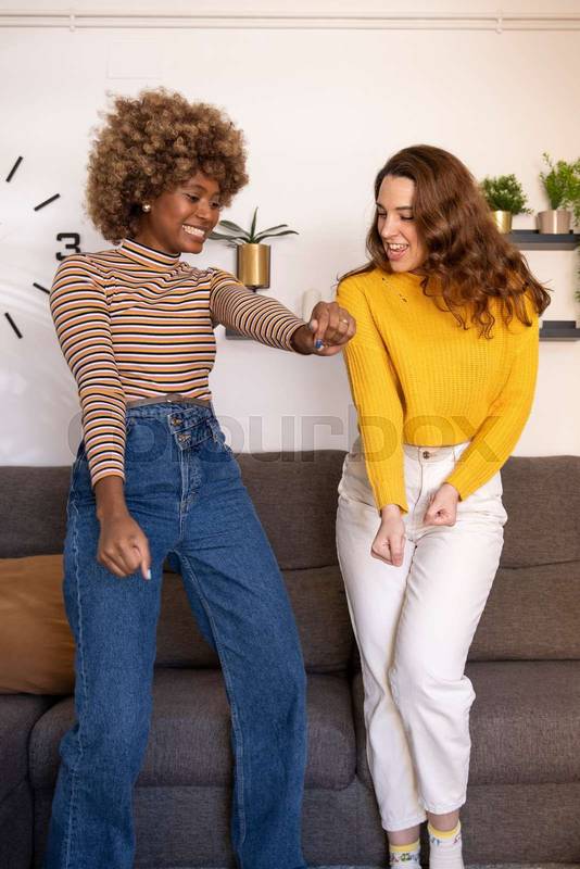 Multiracial young funny women dancing in their apartment. Happy and excited couple of girls celebrating in their living room.