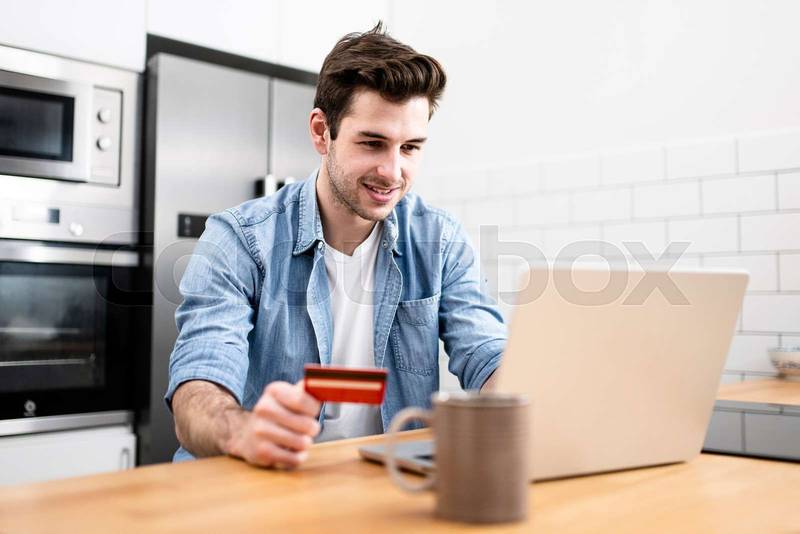 Man sitting in front of the computer holding a credit card at home - Young adult doing a online payment with his laptop at kitchen - business, technology concept