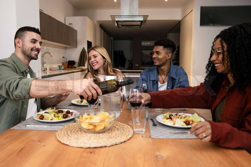 Diverse group of happy friends having party diner at home. Smiling people eating and drinking on modern apartment.
