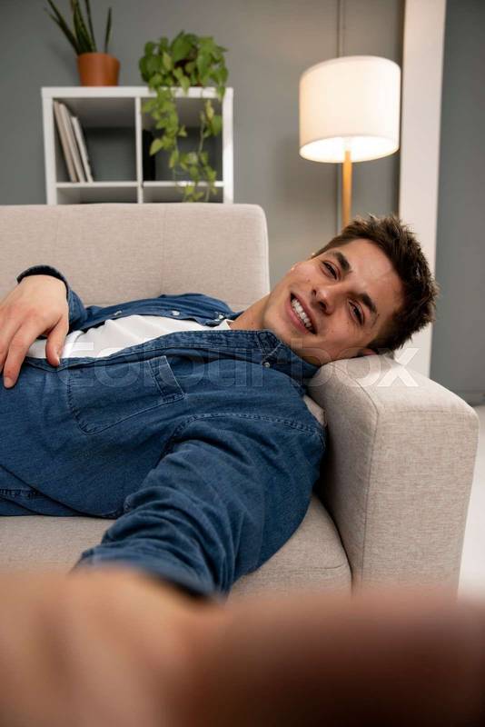 Handsome young adult guy resting at home and taking photo at the living room. Smiling male laying taking selfie while relaxing on home sofa. 