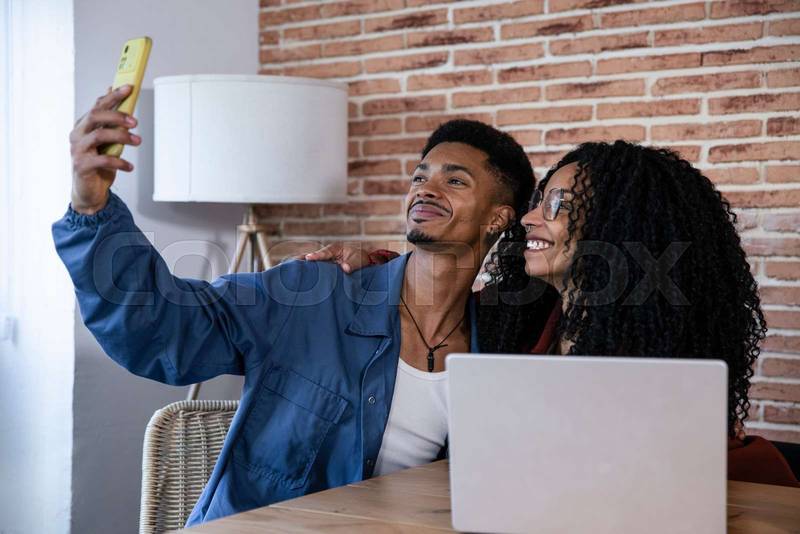 Young couple taking selfie while using computer at home. Young adult friends with laptop and mobile phone taking a picture while sitting on the living room.