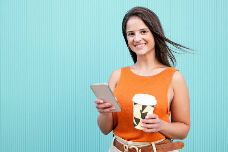 Beatiful woman smiling and looking at camera with a take away coffee and phone in turquoise wall - Portrait of happy young woman smiling and looking at camera