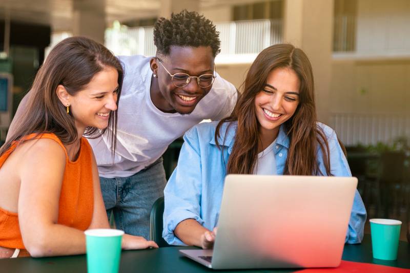 Group of happy friends students sitting in a cafe bar looking at laptop