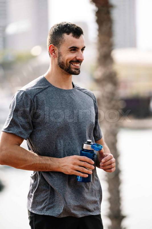 Happy active man drinks and recovery with refreshment after exercise outside. Young sports guy drinking water from bottle walking after workout in a park. 