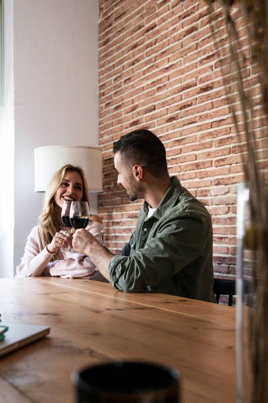 Affectionate couple smiling in a date drinking alcohol sitting at table in a house. Joyful woman and man drinking wine together in a living room with glasses.