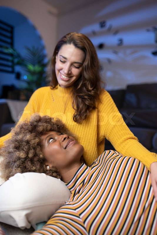 Multiracial couple looking and smiling at each other on sofa. Cheerful lesbian women relaxing and hugging in a living room